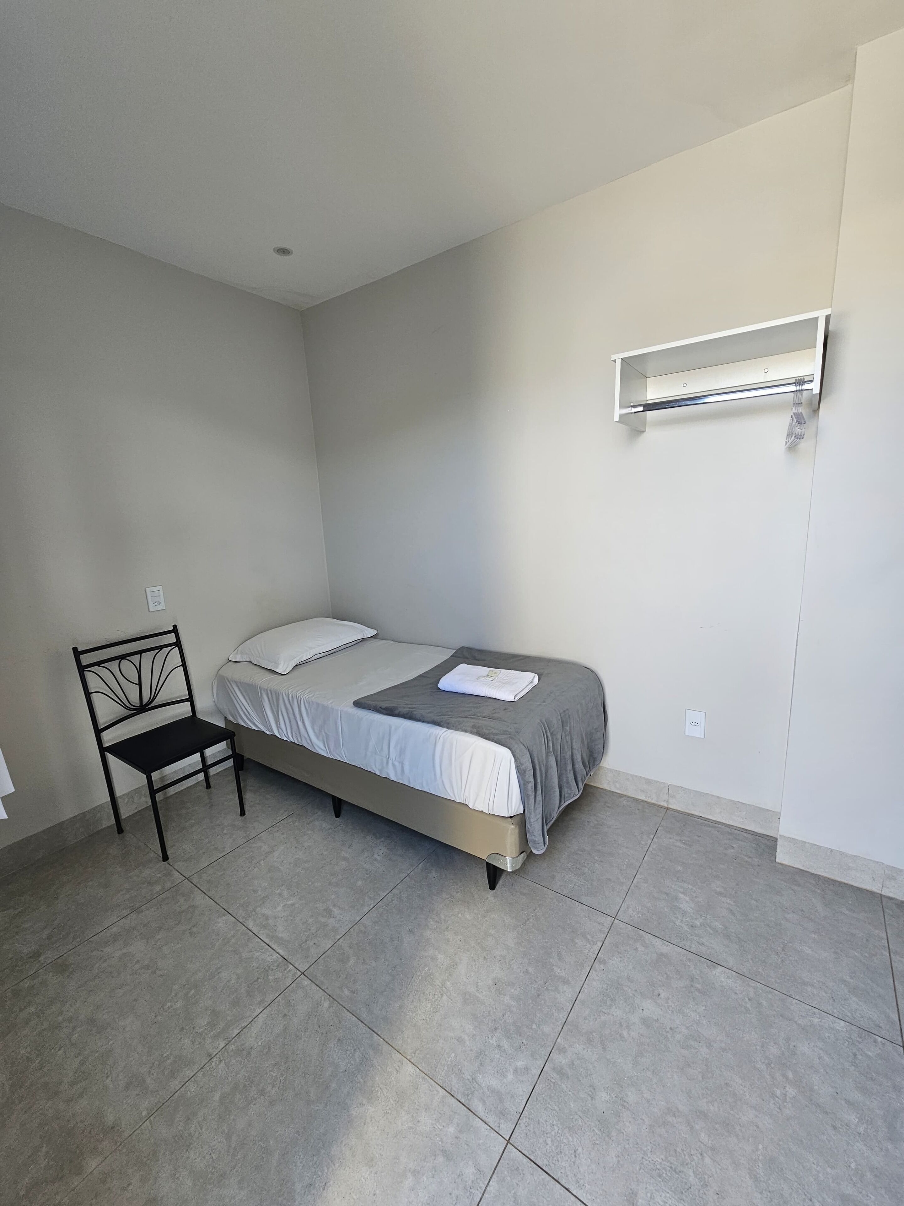 Una habitación pequeña y limpia con una cama individual, una silla y un aire acondicionado de pared.