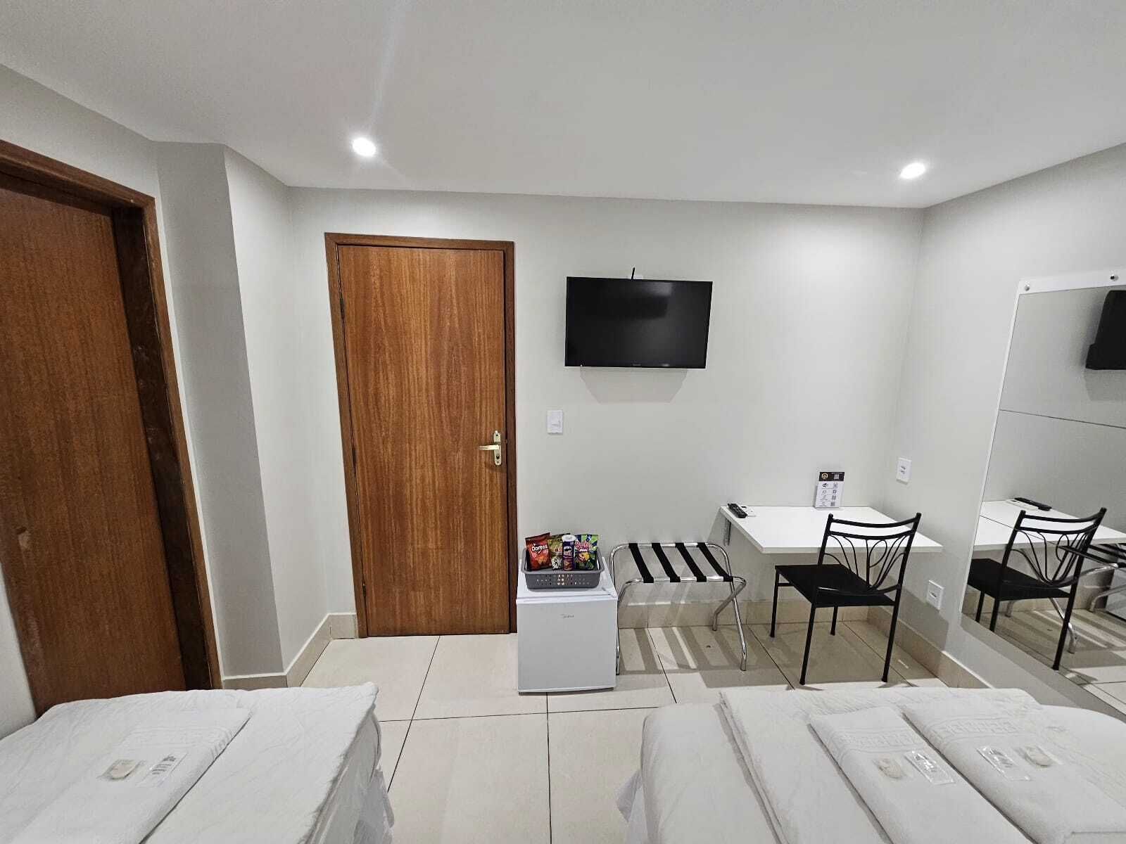 Quarto de hotel compacto com uma cama de solteiro, uma pequena cozinha, uma mesa com cadeira, uma TV na parede e uma porta de madeira.