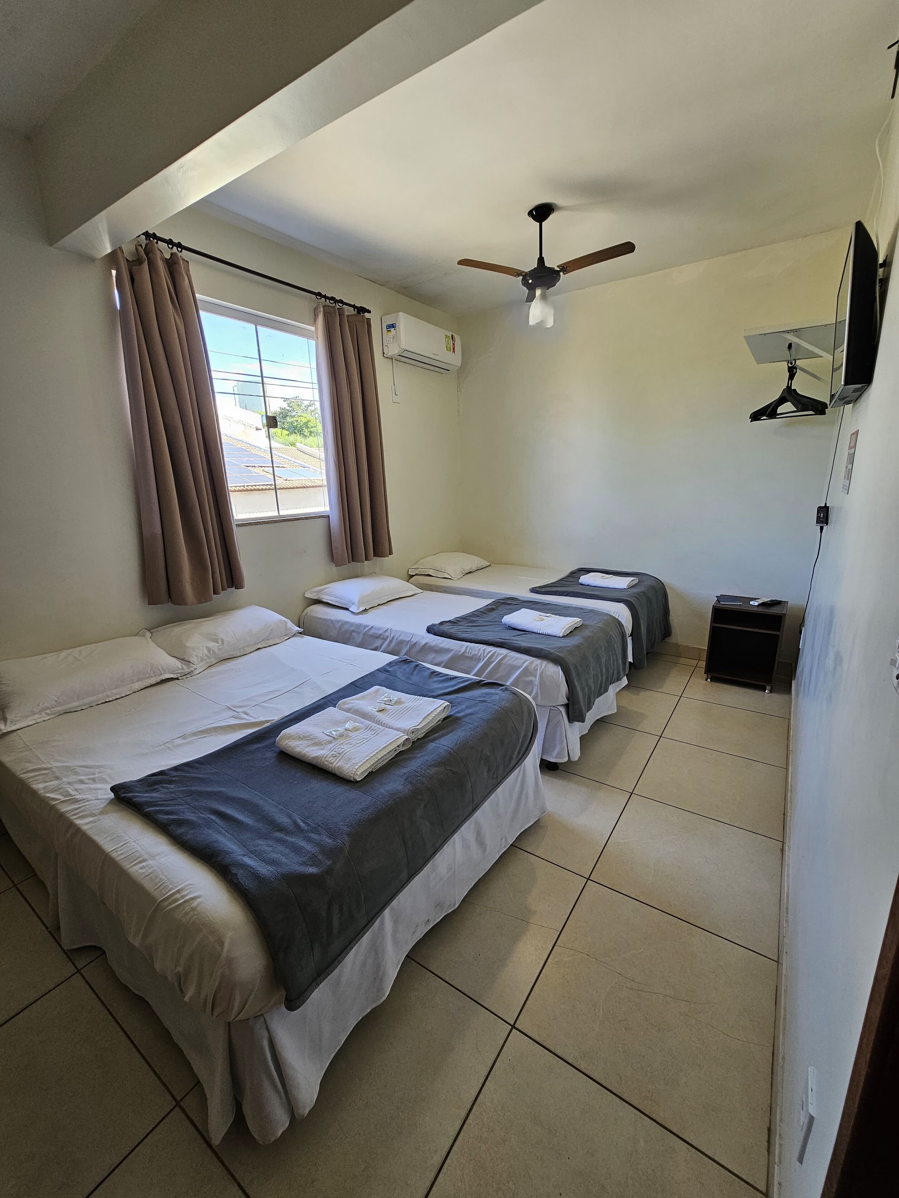 Quarto de hotel com três camas, ar-condicionado, ventilador de teto e janela com cortinas.