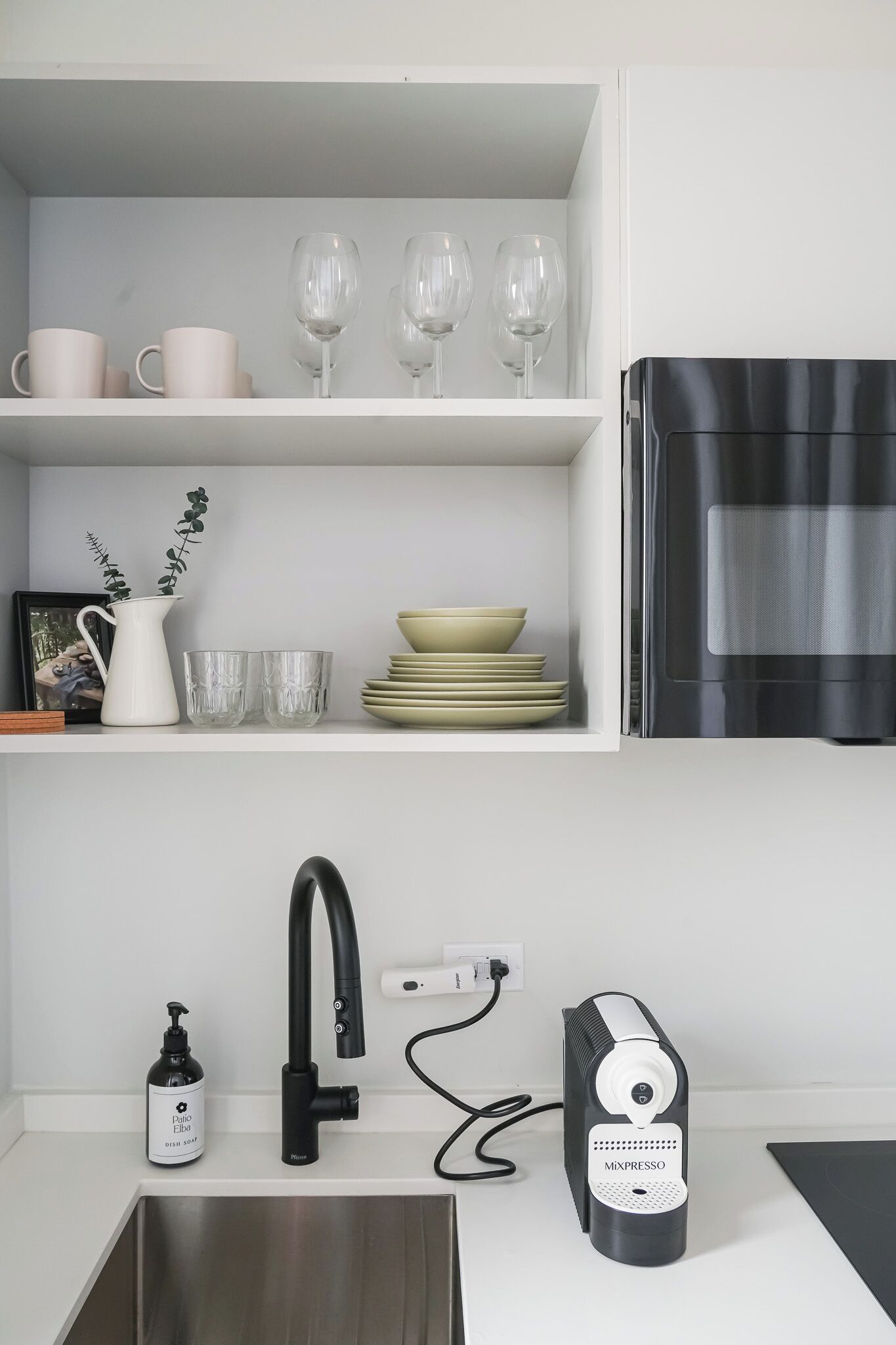 A kitchen with a white countertop, a black faucet, a coffee maker, and a shelf with dishes and glasses.