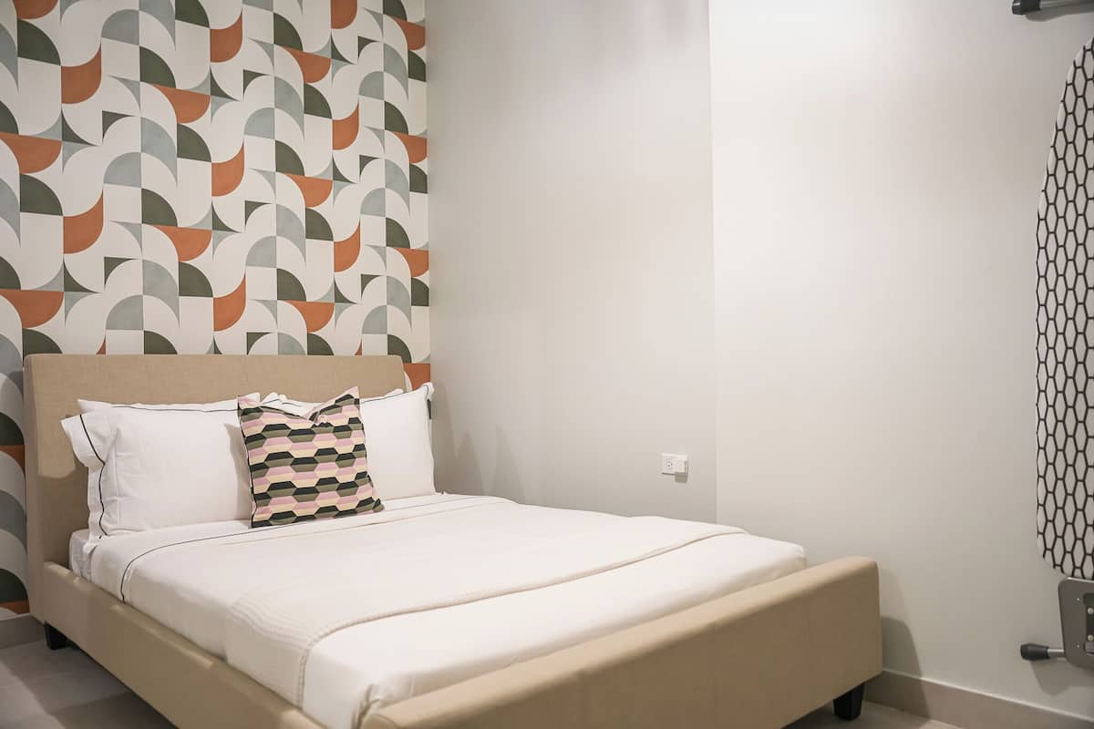 Una cama bien tendida con ropa blanca y un cojín estampado, un cabecero y una pared con diseño geométrico.