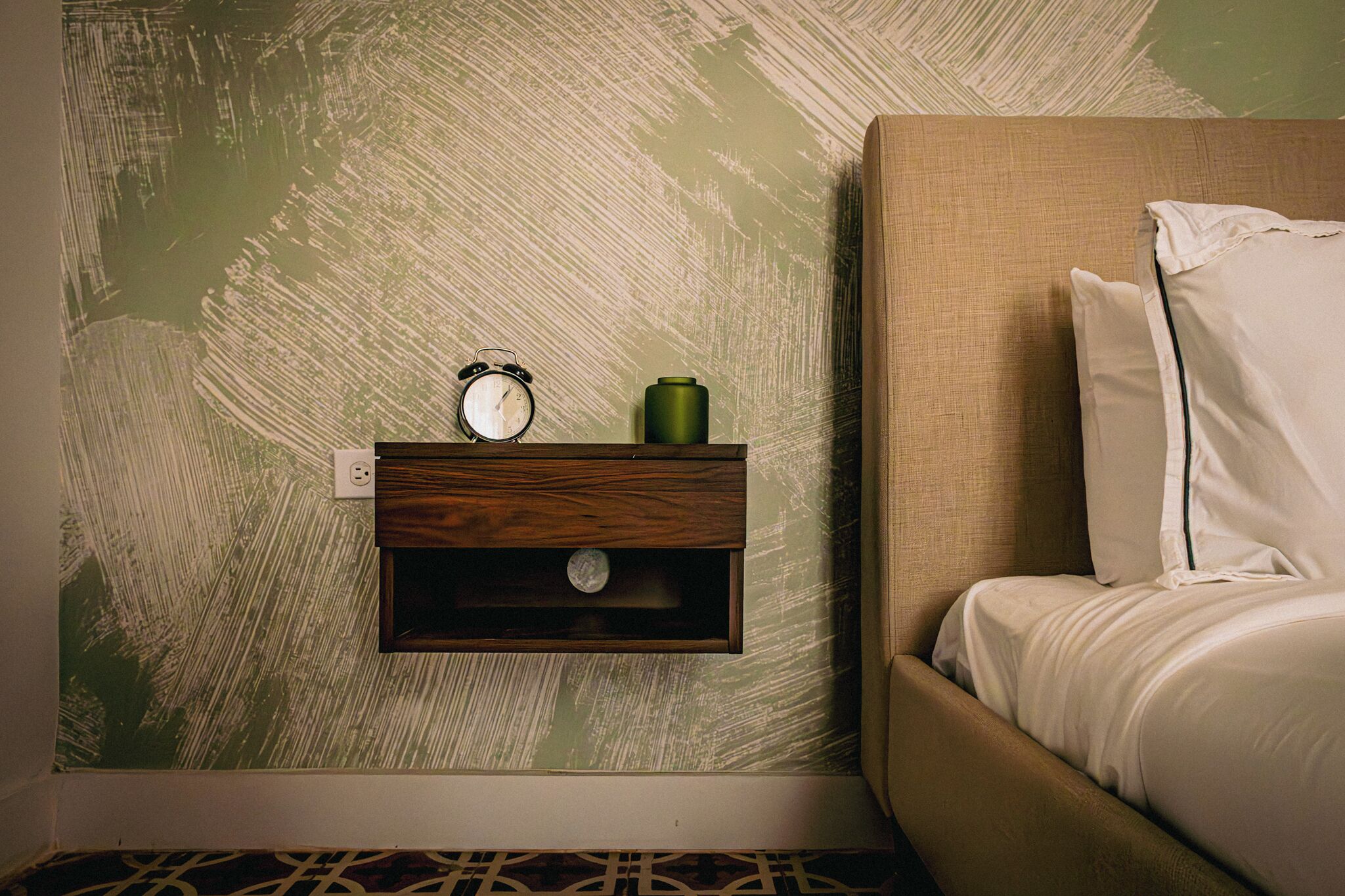 A bedroom with a bed, a nightstand with a clock and a green container, and a textured wall.