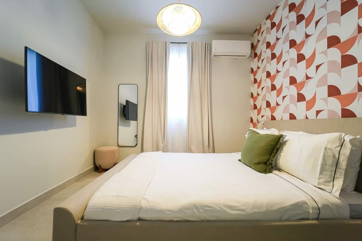 Una habitación de hotel moderna con una cama grande, un televisor de pantalla plana montado en la pared, un reposapiés pequeño y redondo y un papel tapiz estampado.