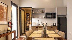 A modern dining area with a wooden table, grey upholstered chairs, and a vase with greenery. A kitchen with white cabinets, a black refrigerator, and a microwave is visible in the background.