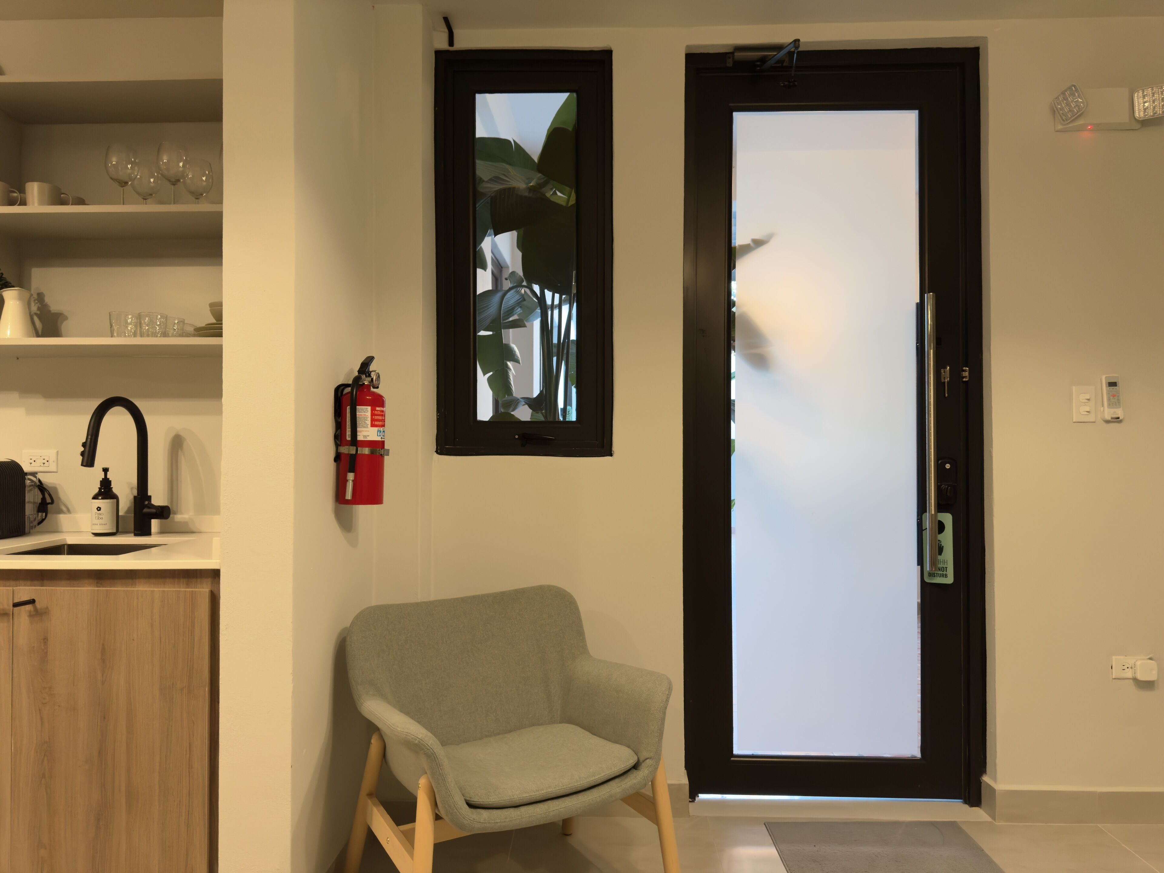 A modern interior with a grey armchair, a kitchen area with a sink and a fire extinguisher, and a door with a frosted glass panel.