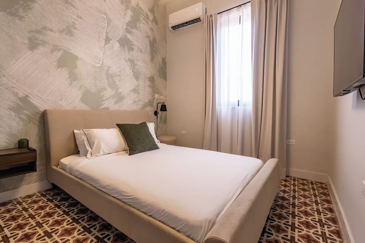 Un dormitorio con cama, mesita de noche, aire acondicionado y ventana con cortinas.