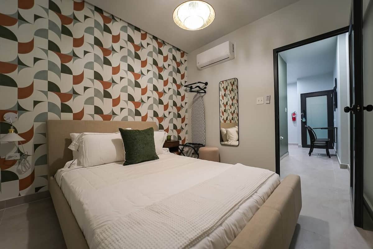 Una habitación de hotel moderna con papel tapiz de diseño geométrico, una cama grande con ropa de cama blanca, una mesita de noche con lámpara, un espejo y una puerta que da a un pasillo.