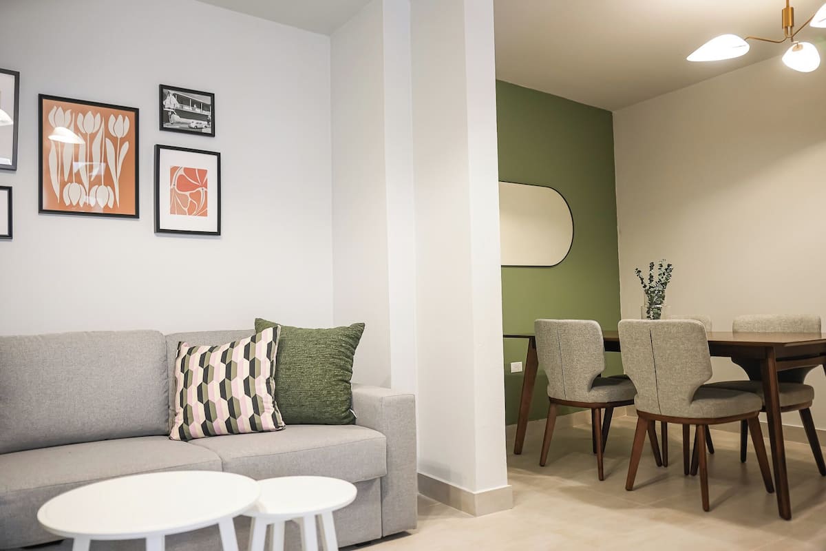 Una sala moderna con un sofá gris, dos mesitas blancas y un comedor con una mesa de madera y cuatro sillas.