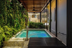 Una piscina con una pérgola de madera en la parte superior, rodeada de abundante vegetación y con vistas a un edificio al fondo.