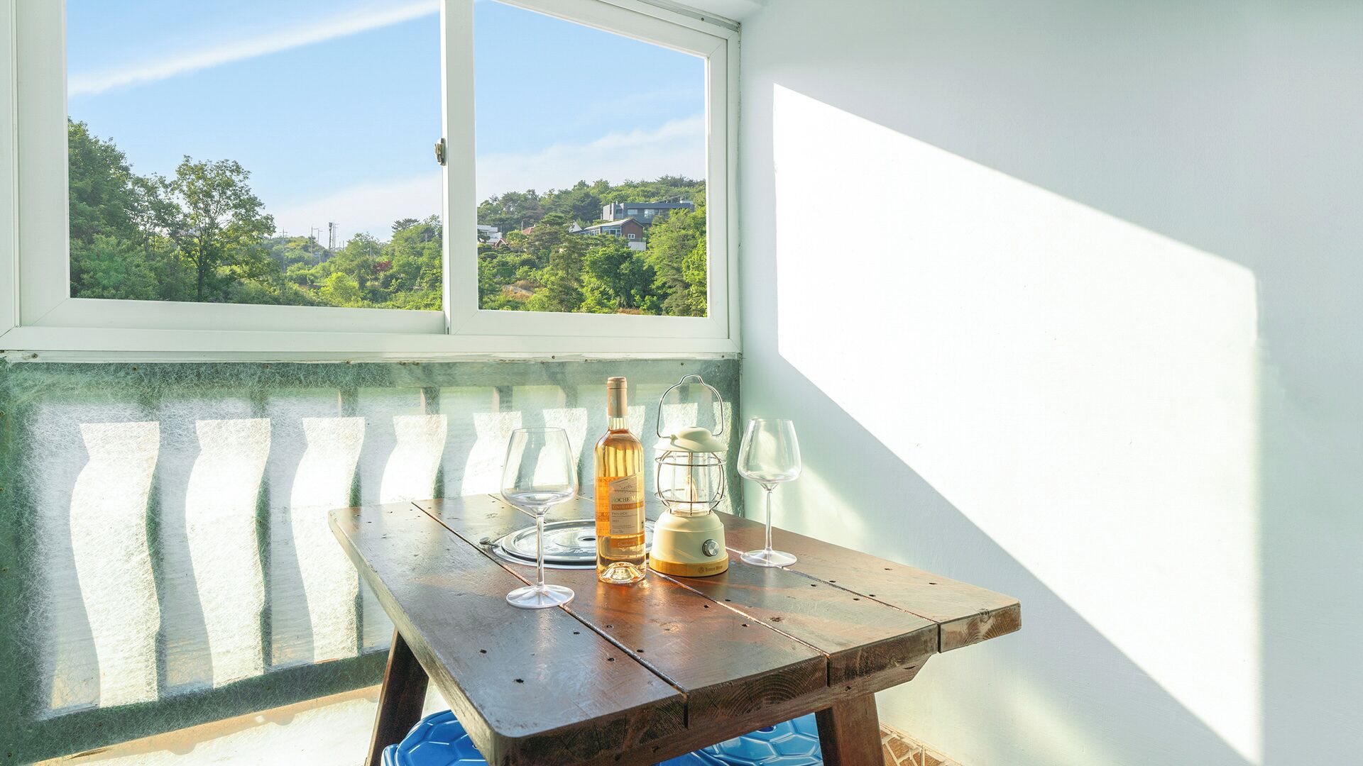 A wooden table with a bottle and two glasses, positioned near a window with a view of greenery and buildings.