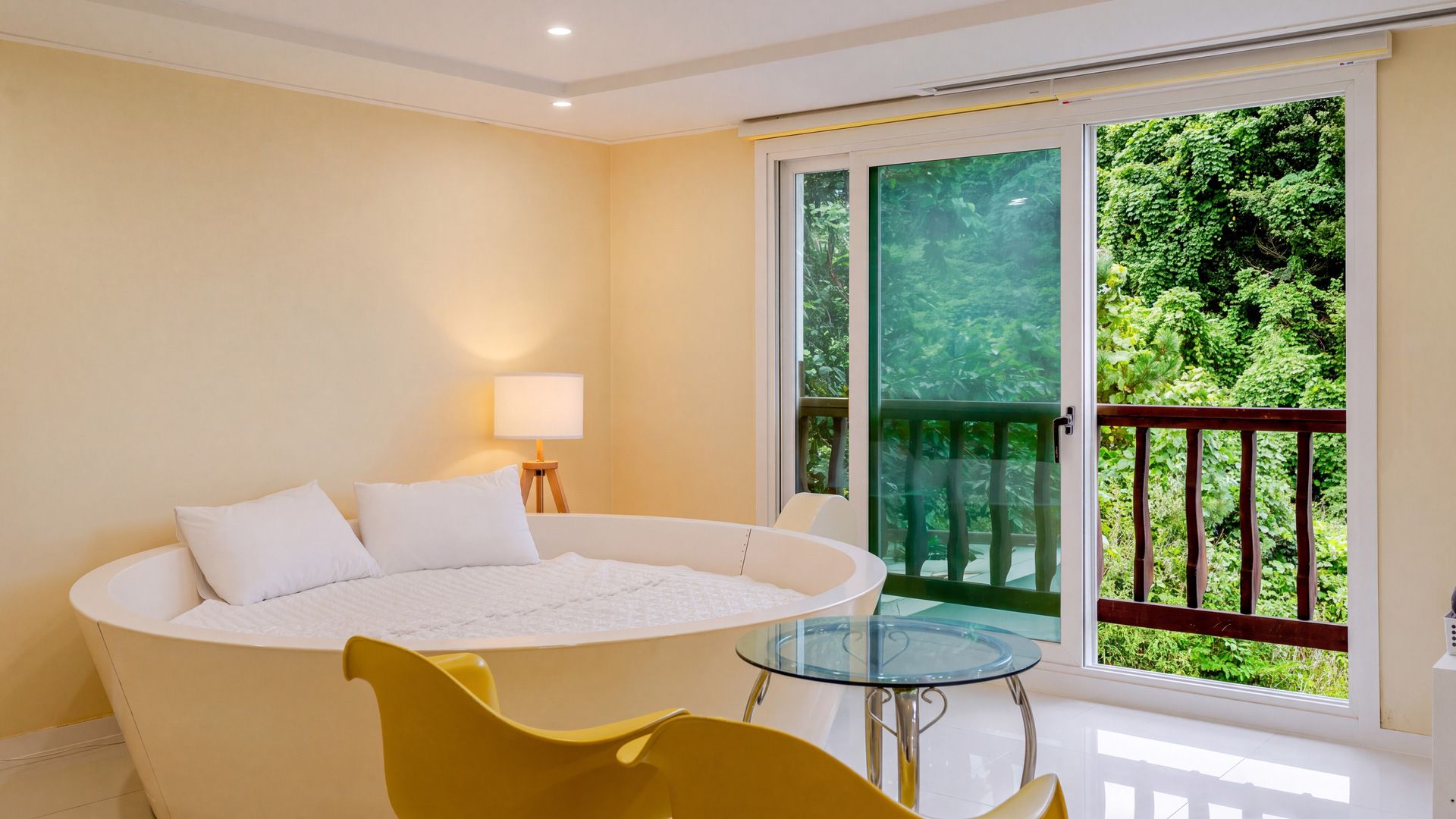 A bedroom with a large bathtub, a glass table, and a balcony with a view of greenery.