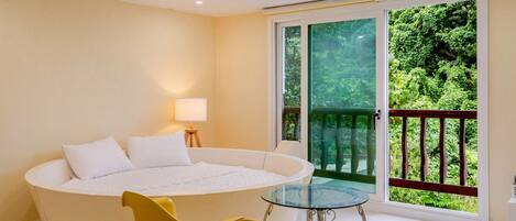 A bedroom with a large bathtub, a glass table, and a balcony with a view of greenery.