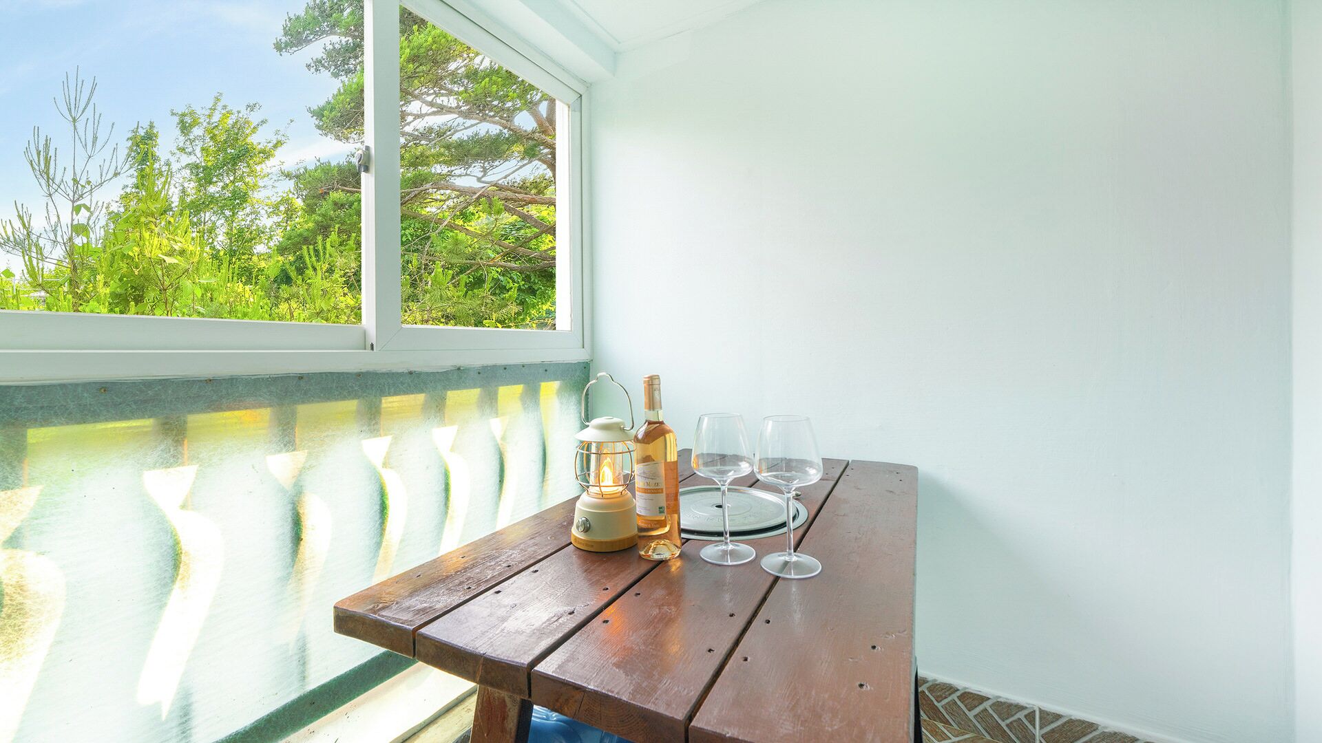 A wooden table with a bottle of wine and glasses, a lantern, and a view of greenery through a window.