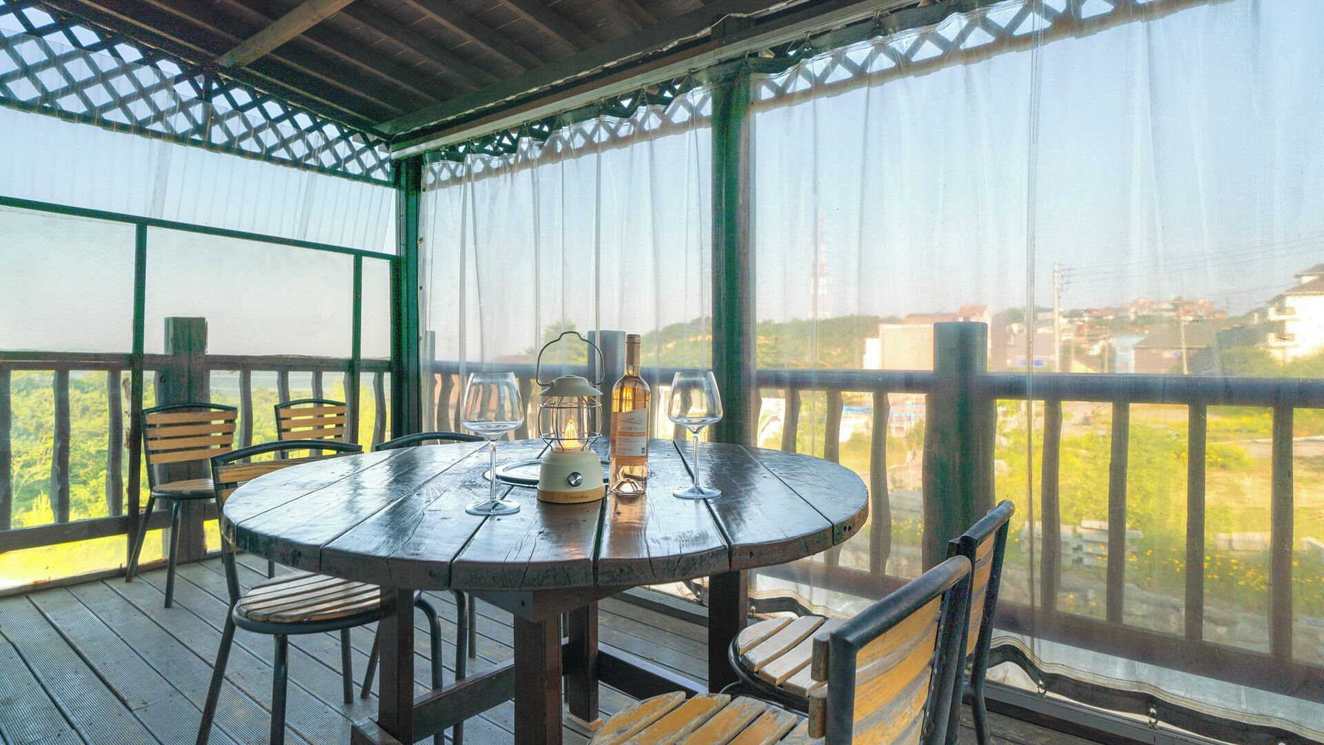 A round wooden table with chairs, a bottle, and a wine glass on a covered outdoor deck with a view of buildings and greenery.