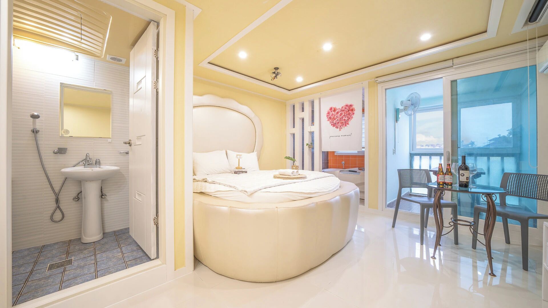 Une chambre à coucher moderne avec un lit aux lignes courbes, une salle de bain avec douche et un balcon équipé d’une table et de chaises.