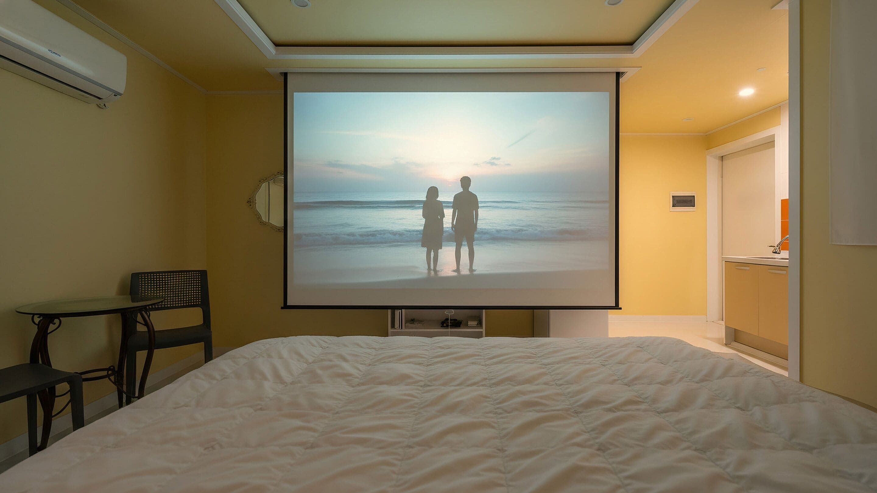 Une chambre comprenant un lit, un bureau, une chaise, un écran de projection diffusant une scène de plage et un coin cuisine.