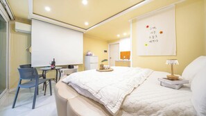 Habitación de hotel con cama, escritorio, silla, mesita con una botella, una planta y un cuadro en la pared.