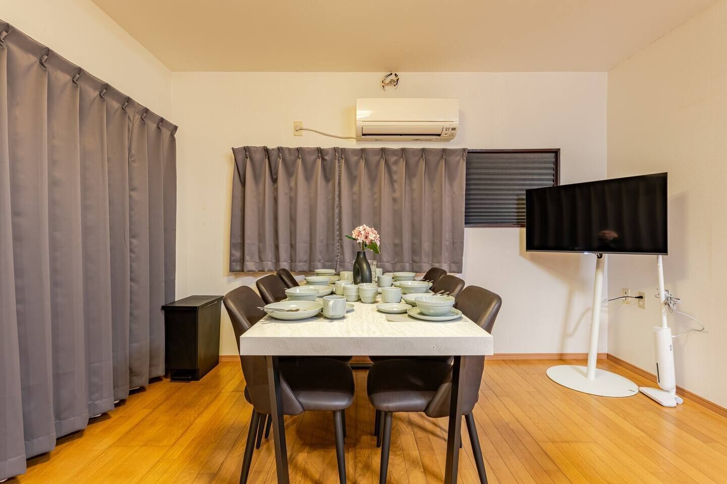 A dining room with a table set for four, grey curtains, an air conditioning unit, and a television on a stand.