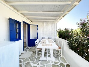 Een overdekt terras met een stenen vloer, een witte tafel en blauwe deuren.