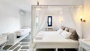 A bedroom with a four-poster bed, a bedside table, a nightstand, a window with curtains, and a view of the outside. - Sandy Side Mykonos by Angels Group (Mykonos)