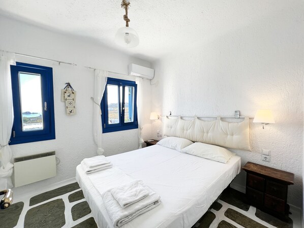 Een hotelkamer met een bed, twee ramen met blauwe kaders, een wit hoofdeinde en een nachtkastje met een lamp.