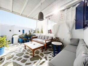 Een zonnig terras met een stenen vloer, een houten tafel, een bank en potplanten.