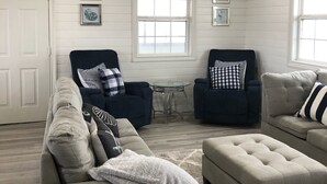 Ein Wohnzimmer mit einem beigen Sofa, zwei Sesseln, einem Couchtisch und einem Deckenventilator. An der Wand hängen drei gerahmte Bilder, ein Fenster und eine Tür.