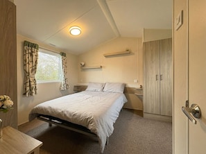 A bedroom with a bed, a window with curtains, a bedside table, a nightstand, a shelf above the bed, and a wardrobe. - Prime lakeside caravan at Haven Golden Sands (Mablethorpe)