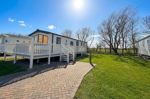 A mobile home with a paved walkway leading to a deck, surrounded by a grassy lawn and trees.