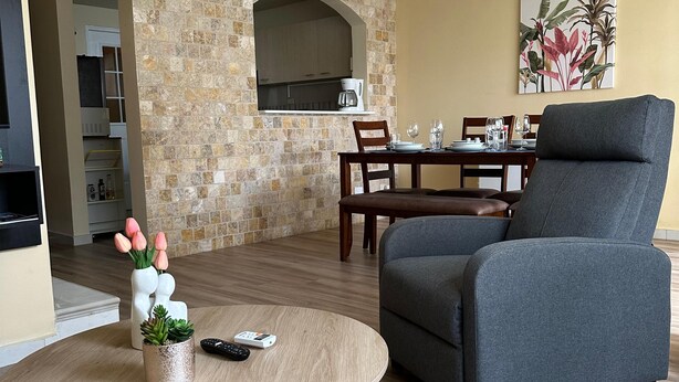 Sala de estar con un sillón gris, una mesa de centro redonda de madera y una pequeña planta en maceta. Hay un comedor con una mesa dispuesta para dos personas, una cocina con salpicadero de piedra y una lámpara colgante.
