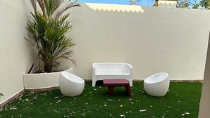 Un pequeño patio con césped artificial, dos sillas blancas, una mesa pequeña y una planta en maceta.