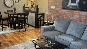 Una sala de estar con un sofá gris, una mesa de centro de madera con escritura, una zona de comedor con un reloj, una araña y una pared de ladrillo con un radio.