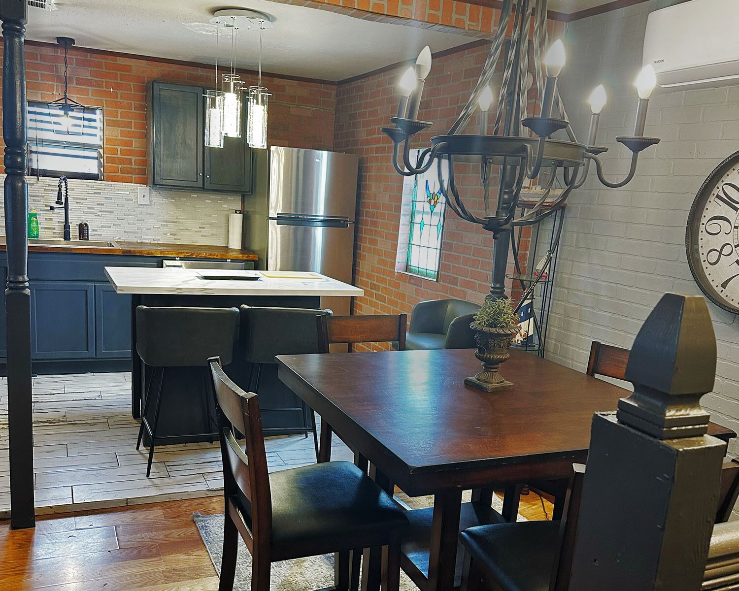A kitchen with a dining area, featuring a chandelier, a clock, and a brick wall.