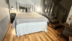 Un dormitorio con cama, suelo de madera, cabecero de metal, mesita de noche y lámpara, además de una escalera.