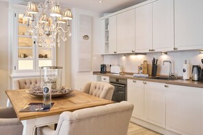 A modern kitchen with white cabinetry, a wooden dining table, and a chandelier. - Domizil am Möwenberg 16 - Baltic Lounge Scharbeutz, Stilvoll Elegante Ferienwohnung in Ruhiger Lage (Scharbeutz)