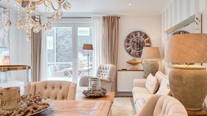 Een ruime woonkamer met een grote houten tafel, comfortabele fauteuils, een kroonluchter en een grote wandklok.