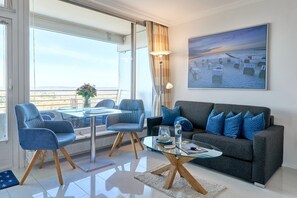 Un salón moderno con una mesa de centro de vidrio, dos sillones azules, un sofá gris con cojines azules y un amplio ventanal con vistas a una playa.