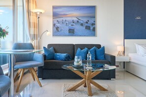 A modern living room with a glass coffee table, a grey sofa with blue cushions, a glass dining table with chairs, and a large beach-themed painting on the wall.