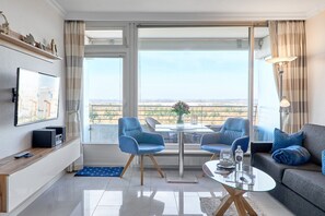 A modern living room with a sofa, two armchairs, a coffee table, and a TV on a stand. There is a balcony with a view of the countryside.