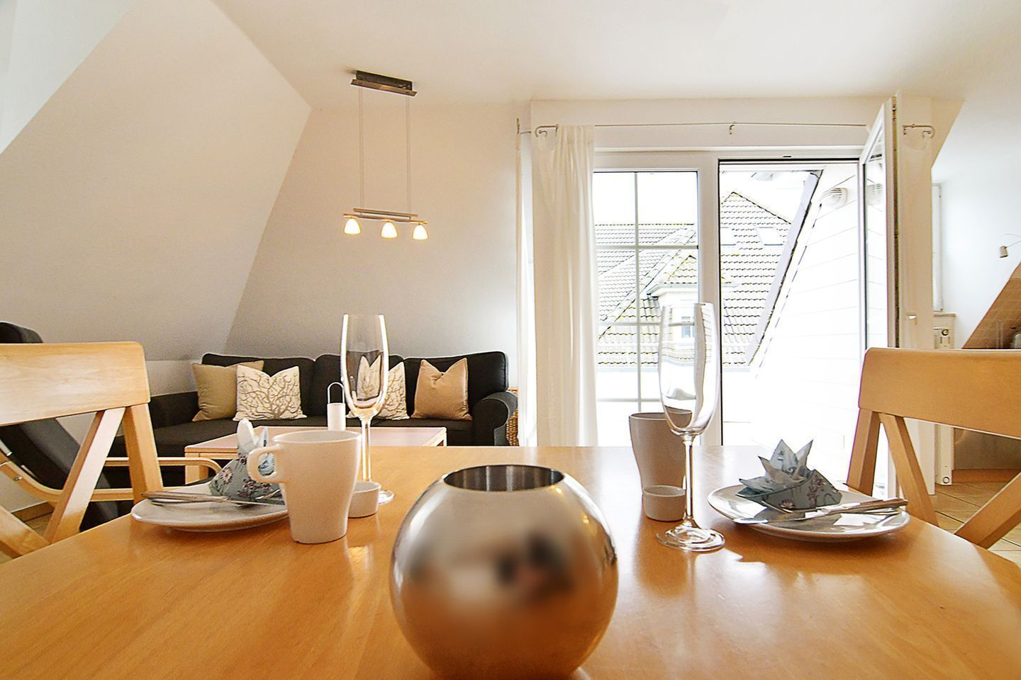 A dining area with a wooden table, chairs, and a modern chandelier. There is a sofa with cushions in the background, and a window with curtains.