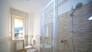 A modern bathroom with a glass shower enclosure, a toilet, a sink, and a window providing a view of the outside. - Vittorio Veneto 9 Int 20 (Borghetto Santo Spirito)