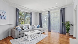 A modern living room with a grey sofa, white coffee table, and a large window with grey curtains. - Sailors Cove | Pet Friendly (Point Frederick)