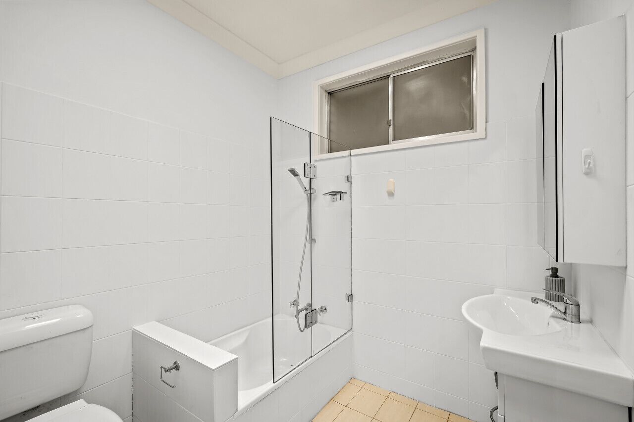 A modern bathroom with white tiles, a glass-enclosed shower, a white toilet, and a sink with a mirror.