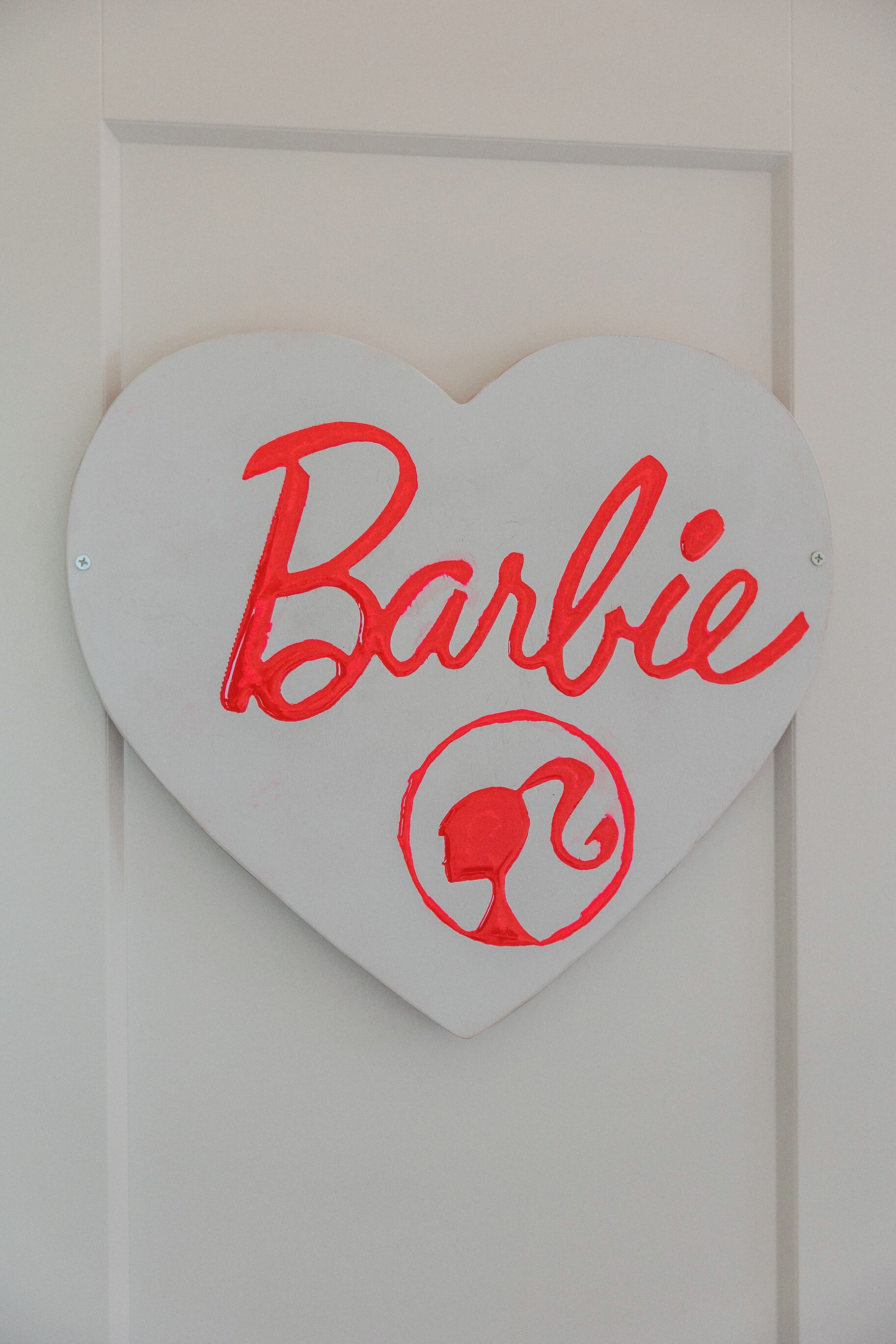 Un letrero blanco con forma de corazón que muestra la palabra 'Barbie' y un logotipo, colocado sobre una puerta blanca.