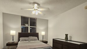 Ein Schlafzimmer mit einem Bett, zwei Nachttischen mit Lampen, einer Kommode, einem Deckenventilator und einem Fenster mit Jalousien.