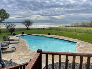 A swimming pool with a stone deck, surrounded by a wooden deck railing, and a view of a lake. - Luxury Lake House with Pool table & Hot Tub (Lakewood Village)