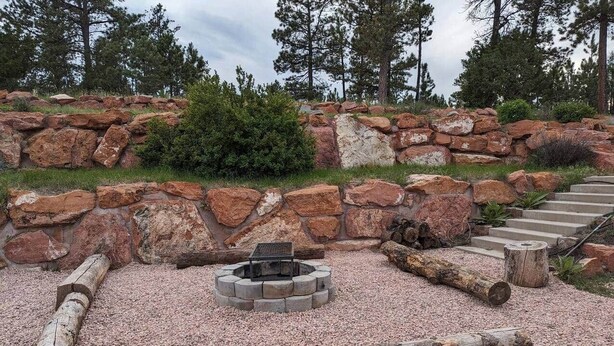 Un hogar de piedra rodeado de troncos y grava, con una escalera que sube a una zona rocosa y árboles al fondo.