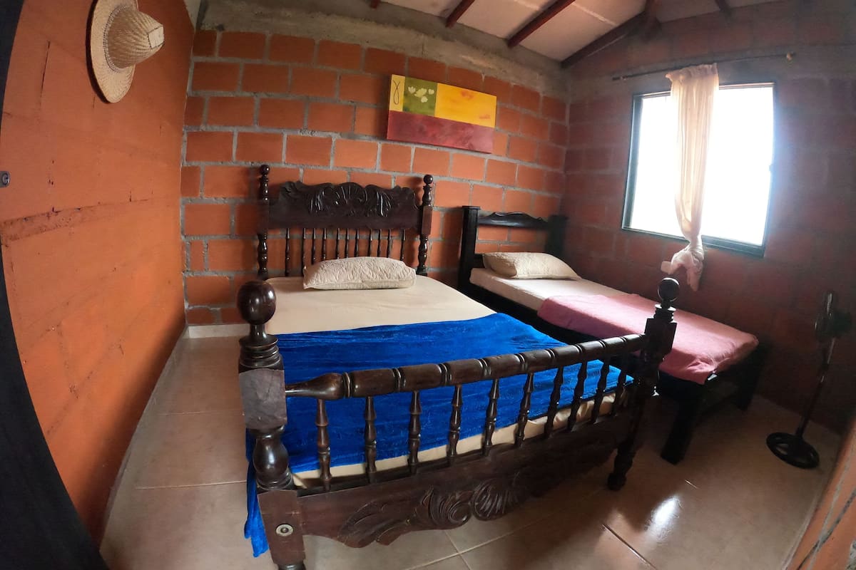 Ein Zimmer mit einem hölzernen Bett, einem kleinen Tisch, einem Stuhl, einem Ventilator und einem Fenster mit Vorhängen.