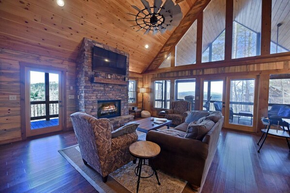 A log cabin interior with a stone fireplace, wooden floors, and large windows offering views of the outdoors.