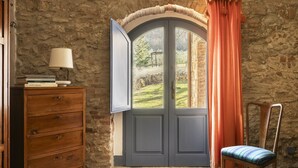 Une pièce avec un mur en pierre, une commode en bois surmontée d’une lampe, une chaise et une fenêtre avec des rideaux.